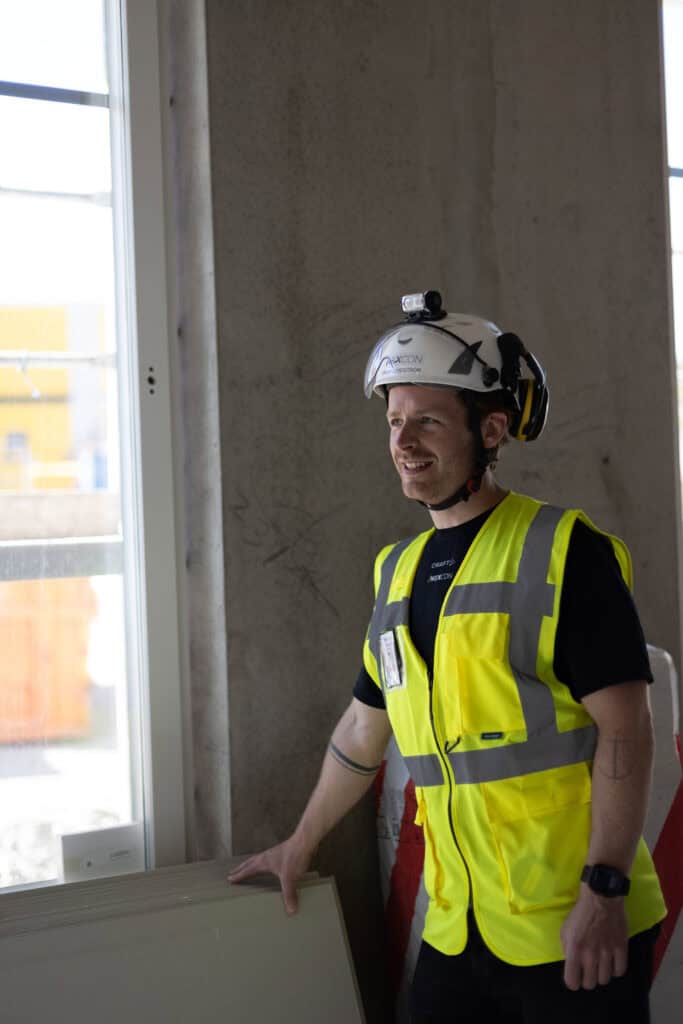 Byggarbetare i hjälm och reflexväst som arbetar inomhus, står vid ett fönster i ett byggprojekt i Up.