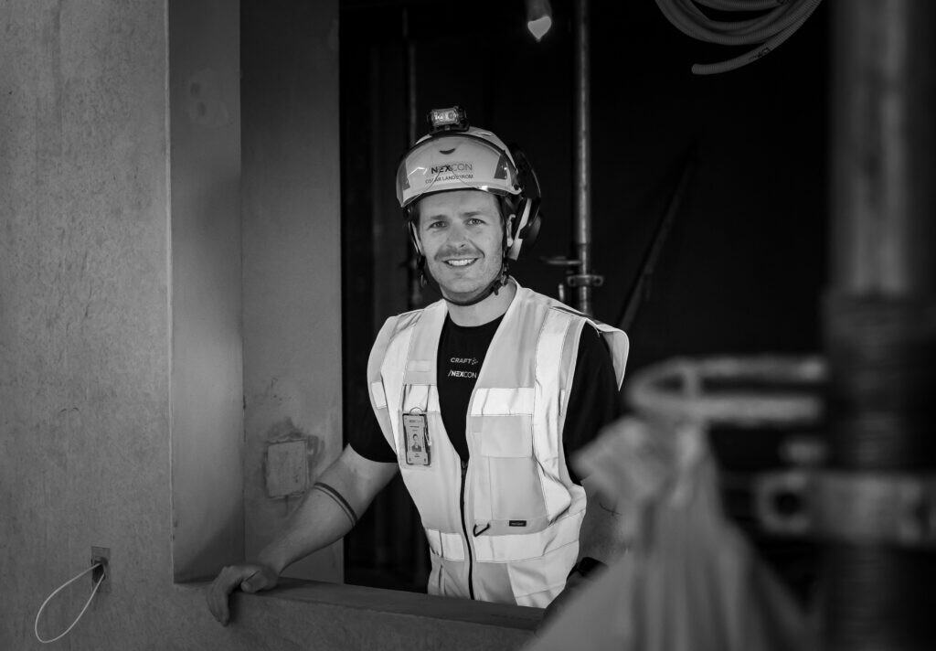 Arbetsbild av en byggarbetare i arbetskläder och hjälm, leende och ser glad ut.