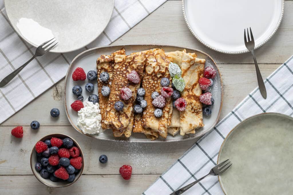 Pannkakor med bär, grädde och serveringsfat på träbord.